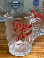 Close-up of a clear glass beer mug with handle featuring a red 'Beef Baron' logo and illustration of a woman, with address text '624 York St., London, Canada' below the logo.