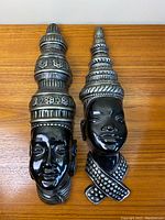 Two black chalkware wall hangings of African women with silver headdresses and necklaces shown side by side on a wooden surface.