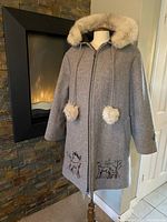 Front view of the gray wool winter coat on a mannequin, showing fur-lined hood, drawstrings with fur pom-poms, front zipper, and embroidered deer design near the hem.