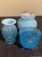 Three blue glass items including hobnail vase, rose vase with clear ruffled rim, and EAPG tri-footed dish all on dark surface