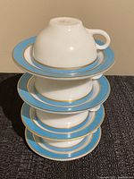 Stacked display of four white milk glass tea cups each sitting on matching turquoise blue and gold trimmed saucers.