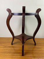 Front view of vintage walnut plant stand showing three curved legs, the round upper frame, and a triangular lower shelf with visible repair marks on the legs.