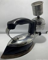 Photo of antique metal iron with black handle and cylindrical Coleman-branded fuel reservoir at the back, top-down angle.