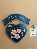 Heart-shaped welcome wall hanging painted blue with pink and white flowers and 'WELCOME' text on a ribbon banner above, size approximately 9.5 by 9 inches.
