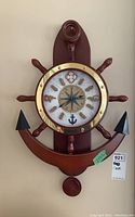 Front view of the nautical themed wooden wall clock showing the anchor shaped wooden base, ship wheel handles, and brass circular clock frame with nautical elements on the face.