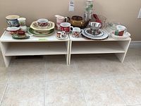 Photo showing two low white shelves topped with assorted dishes including mugs, plates, teapot, and tumblers