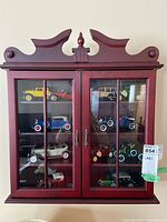 Front view of the red wood display case mounted on the wall, glass doors closed, showing multiple collectible toy cars inside.