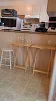 Photo of two matching folding wood tables and a painted wooden stool positioned in front of kitchen counter with glassware on tables (not included).