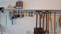 Wall-mounted garden tools including fan rake, edger, spades, and potato fork with wooden handles, along with painting brushes on hooks