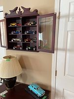 Wall-mounted wooden display case with 11 smaller model cars in varying vintage styles and colors.