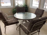 Patio set arranged in a bright indoor area showing 4 cushioned metal frame patio chairs around a round glass top table with metal frame.