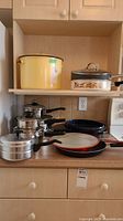 Display of assorted pots and pans including yellow enamelware stock pot, sauce pots, roasting pan, frying pans on kitchen counter and shelf