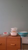 Full view of stacked CorningWare white casserole dishes with lids showing floral pattern, alongside turquoise blue Pyrex mixing bowl with white patterns.