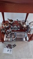 Full view of the entire silver plate tea and coffee service set on wooden lower shelf, showing teapot, creamer, sugar bowl, salt and pepper shakers, stainless steel toast rack on tray, small stainless cup, and additional pot.