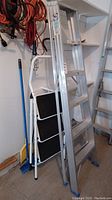 Two step ladders: an aluminum ladder leaning against the wall and a white kitchen step ladder with black steps next to it.