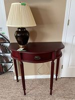 Demi-lune style table with dark mahogany finish, showing overall front view with ceramic black lamp on top and pleated beige lampshade. Table has one oval drawer with decorative pull.