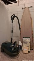 Photo showing vacuum, ironing board, iron, vacuum and dust bags, along with black clothes hangers hanging above