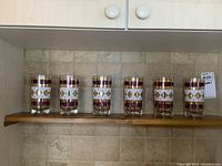 Set of 6 vintage drinking glasses arranged on a wooden shelf with a tiled background, showing their size and decoration.