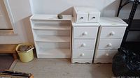 Three white wood storage furniture pieces: two 3-drawer stands, one small 2-drawer unit, and part of a 3-shelf bookcase. A black bag and yellow bucket nearby.