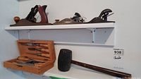 Photo showing three hand planes on a white shelf above a wooden box containing chisels, with a black wooden mallet laying horizontally below the shelf.