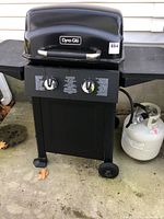Full view of the Dyna-Glo gas barbecue grill with propane tank connected, standing on concrete outside a house.