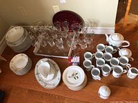 Overview of dinnerware and glassware including plates, bowls, mugs, coffee pot, creamer, sugar, silver trays, and wine glasses on wooden floor.