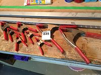Eight small metal hand clamps with red cushioned handles and jaws arranged on wooden surface.