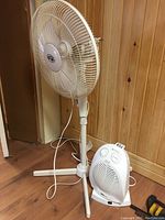 White Hampton Bay pedestal fan with three-leg base and white power cord, positioned against a wooden paneled wall.