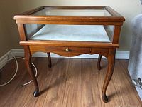 Front view of the glass display table showing wood frame, glass top, curved legs, and small drawer underneath.