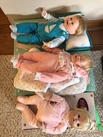 Three dolls arranged side by side, one wearing teal overalls, one in peach outfit, and one in pink romper.