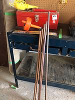 Red metal toolbox sitting on blue metal shelf with several copper pipes leaning against it and an orange drain snake on the floor in front.