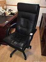 Black leather office chair positioned beside wooden desks in an indoor office setting. Shows front view emphasizing high backrest and padded seat.