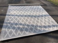 Full view of the outdoor rug partially rolled up on a driveway showing the entire size and diamond pattern.