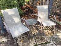 Two outdoor swing chairs with metal frames and gray mesh fabric seat and backs, positioned on stone outdoor patio with occasional small round glass-top table between them.