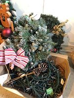Box with mixed small wreaths, pine cones, red striped bow, and coiled green string lights.