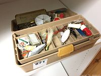 Beige tackle box open showing compartments filled with assorted fishing lures, hooks, and accessories.