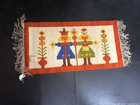 Front view of the carpet showing folk art figures and floral designs, bordered in orange with fringes at each end.