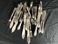 Overview of the entire set of ornate silver fish knives and forks laid out on black fabric, showing the design and condition of the pieces.