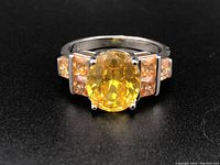 Front view of silver ring with large oval yellow gemstone center, flanked by four smaller square yellow gemstones on each side.