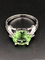 Front and top view of the silver ring with a large oval green gemstone held by prongs, showing detailed band design with small clear accent stones.