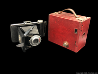Full view of the vintage foldable camera beside red carrying case showing overall items included in lot.