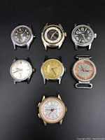 Top-down photo showing all seven vintage watch cases arranged on a black surface, highlighting their different dial designs and wear.