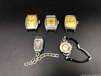 Group of five Deco style vintage watches on black background, showing various dial shapes and metal cases including one with metal link band and one with black wrist strap.