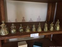 Full set of nine brass figurine bells displayed on a wooden shelf, each uniquely shaped as a female figure in period attire.