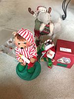 Annalee elf figurine wearing red and white striped nightcap holding a candle, plush moose with Christmas scarf and Santa hat, small Santa Claus figurine, and red Hallmark box.