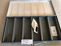 Photo of the metal storage case with numbered slots designed to hold photographic slides, showing some empty slots and a few slides inserted.