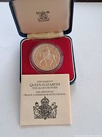 Silver Jubilee proof coin in protective capsule inside red velvet interior case with cream lining and royal crest, piece accompanied by official certificate for Queen Mother 80th birthday commemorative crown.