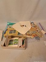 Wide photo showing a box and plastic bag containing assorted postage stamps including loose stamps, envelopes with stamps, and some sheets