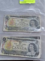 Front view of two Canadian one dollar banknotes in plastic sleeves, showing portraits of Queen Elizabeth II and serial numbers.