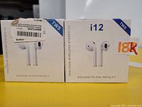 Photo showing two i12 TWS wireless earbud boxes, white color, labeled with 18K sticker on one box. Each box shows an image of the earbuds and text 'Auto power-On Auto Pairing 5.0'.